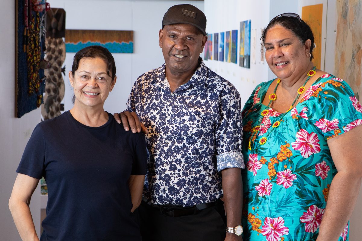 The judging panel of the 2023 Gab Titui Indigenous Arts Award is standing side by side and smiling at the camera. On the left is judge Avril Quaill, who is a woman dark brown hair pulled back and a navy blue shirt. In the middle is cultural advisor John Armitage, who is a man wearing a black cap, navy blue and white floral button down and black pants. On the right is cultural advisor Flora Warrior who is a woman with curly black hair tied back, sunglasses on her head and a bright turquoise dress with pink and orange flowers.