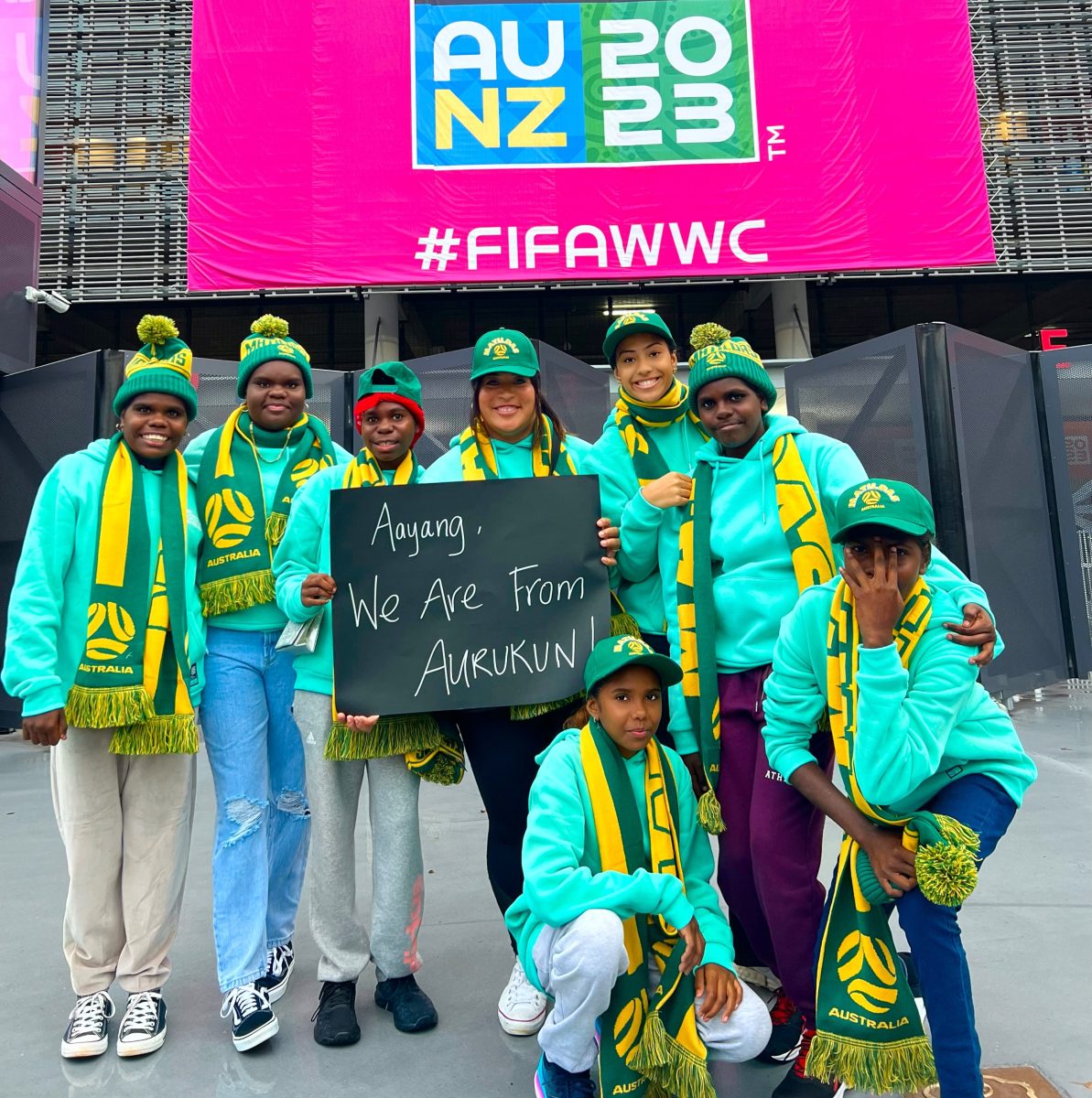 Eight women are posing in front of the FIFA Women's World Cup stadium. The two women in the middle are holding a black sign that reads "aayang, we are from Aurukun" in white handwriting.