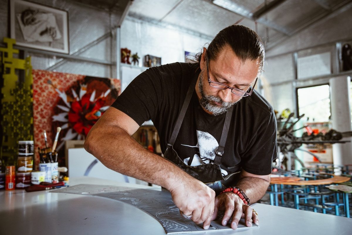 Brian Robinson is a man with dark brown hair pulled back, a black t-shirt, black apron and rectangular glasses. He is carving out designs on a silvery grey sheet placed on a white workbench.