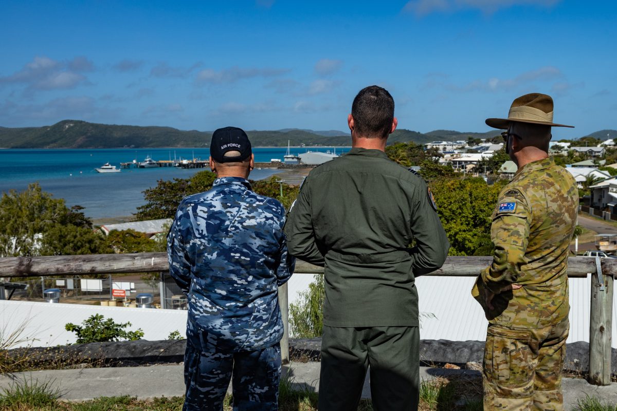 Three men wearing an Australian Air Force uniform, 35 Squadron uniform and camouflage army uniform are standing side by side looking out at the coast with their backs to the camera