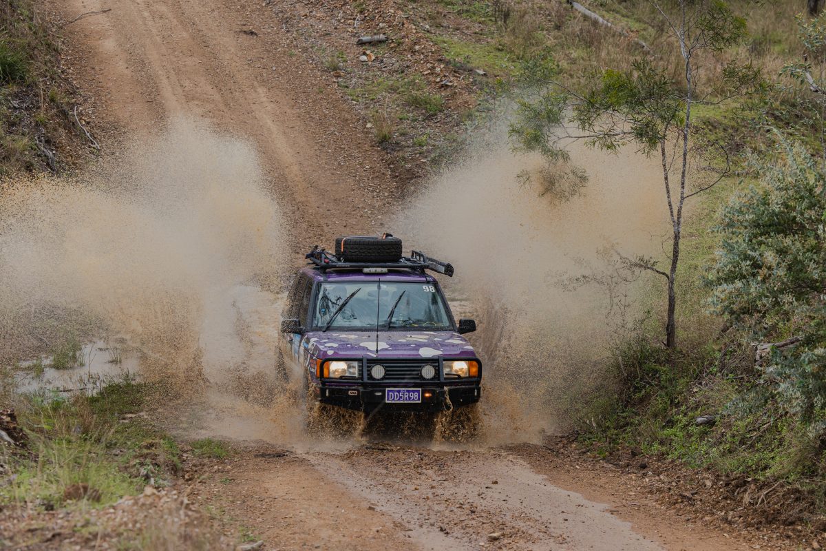 A purple car has made a big splash on a wet dirt road after driving down a hill.