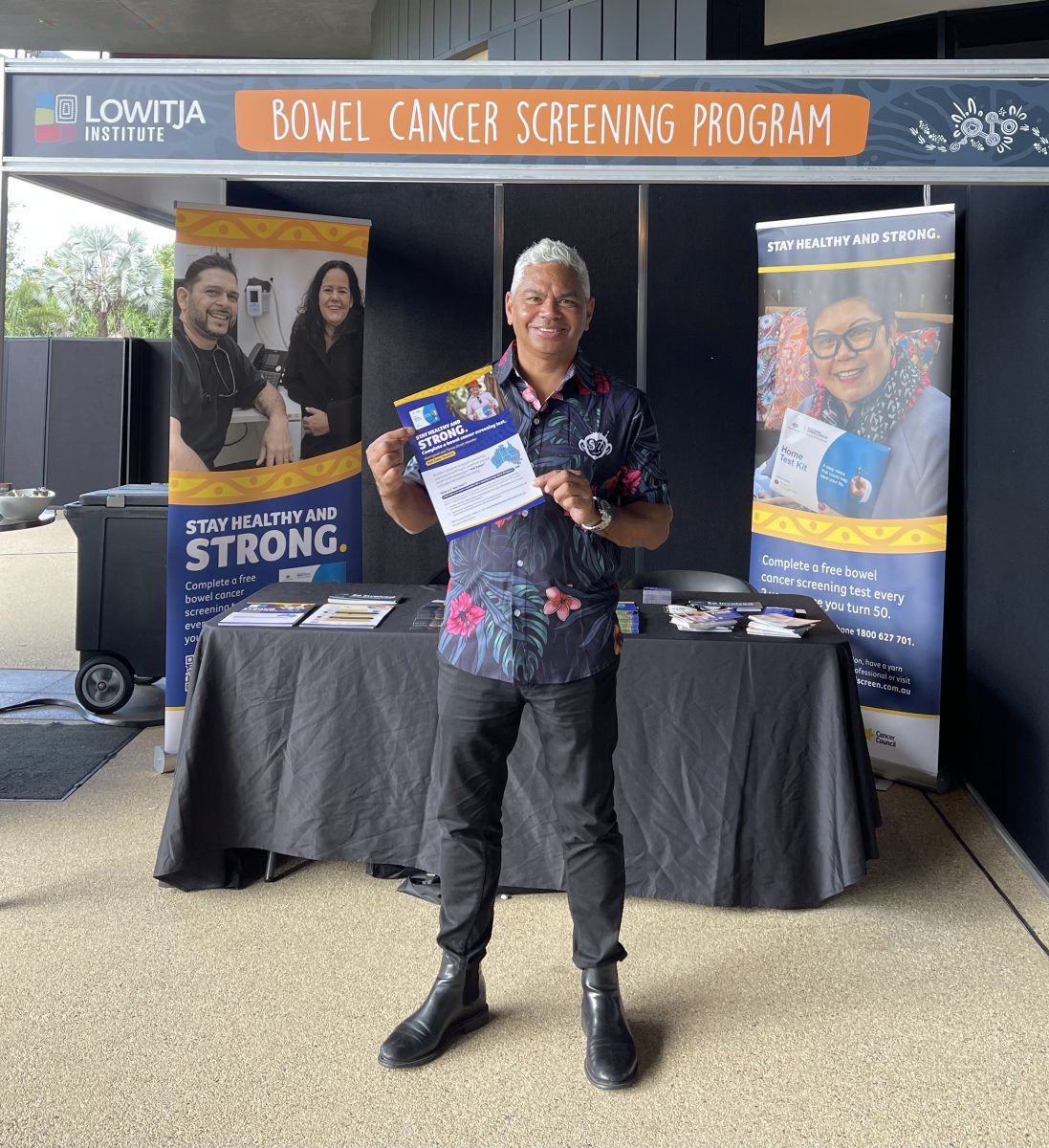 John Paul Janke with white hair, a navy blue button down with pink and orange flowers, black pants and black boots. He is standing in front of a bowel cancer screening program outdoor stall setup, and is smiling at the camera holding a bowel cancer screening flyer.