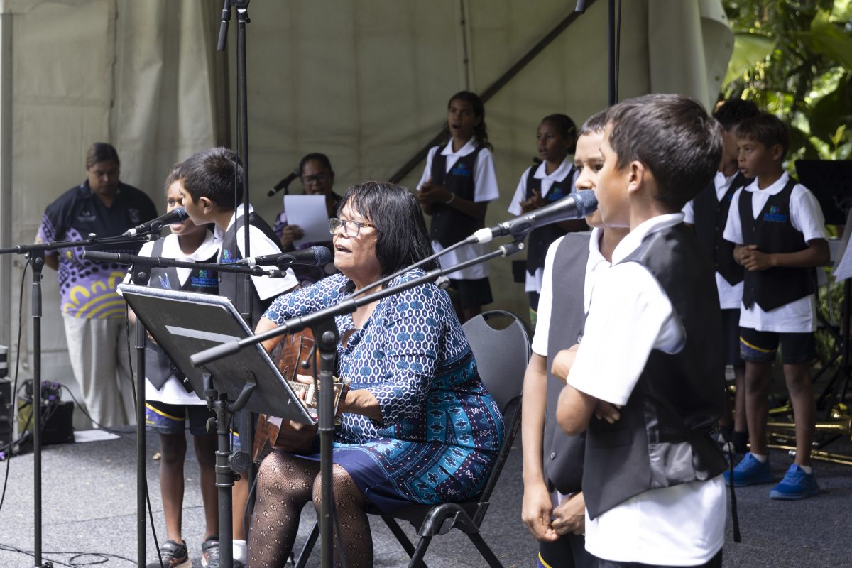 Side view photo of the front of the stage showing Auntie Dora sitting with 4 students either side of her, two on each microphone.