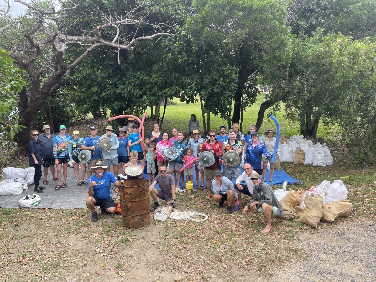 A group photo with participants holding up items found up the main items found on the clean up such as echo sound satellite buoys and pool noodles. In the background are multiple bags of rubbish.