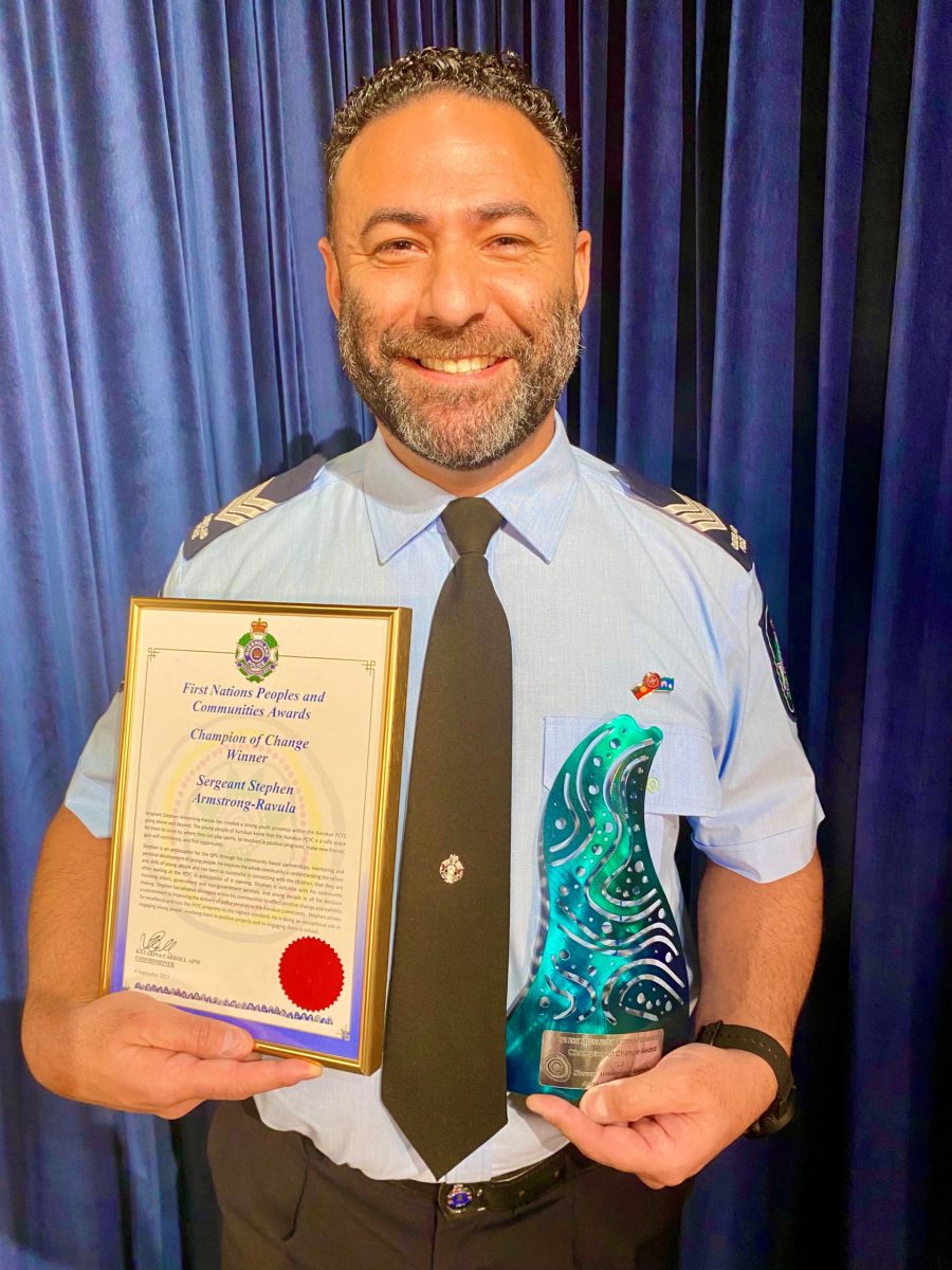 Sergeant Stephen Armstrong-Ravula is a man with short brown hair, moustache and beard. He is wearing a police uniform and is holding a certificate in one hand an a small blue trophy in the other.