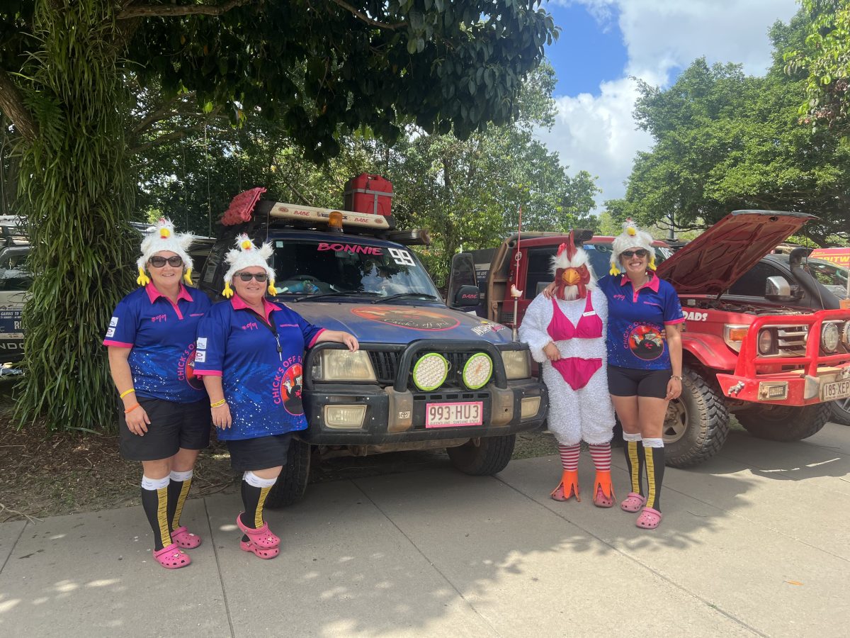 A rally team dressed in blue and pink smile in front of their blue 4WD with pink accents. Three of them are wearing rooster hats, while has on a rooster costume complete with a pink bikini over the top.