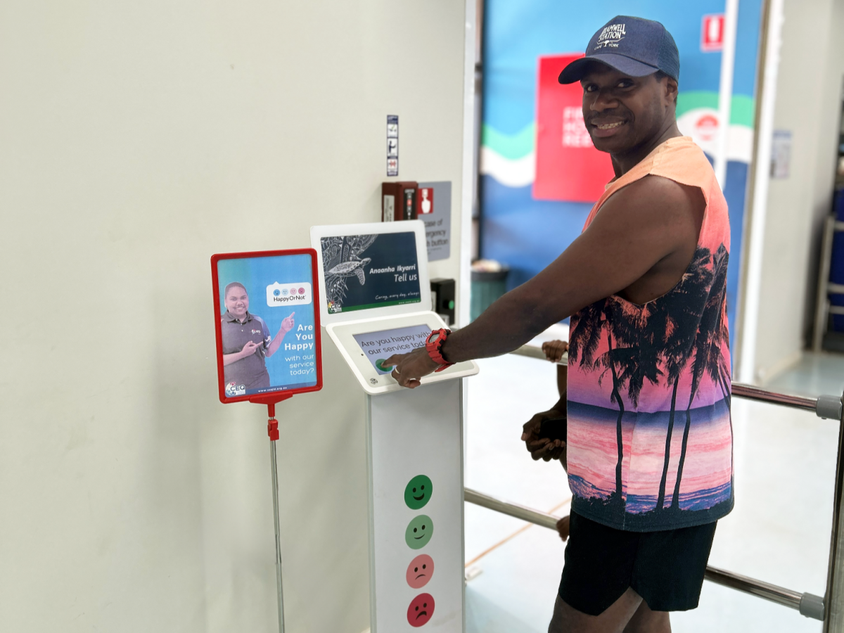 A man wearing a blue cap and a coral short with palm trees is smiling at the camera while using the customer feedback kiosk.