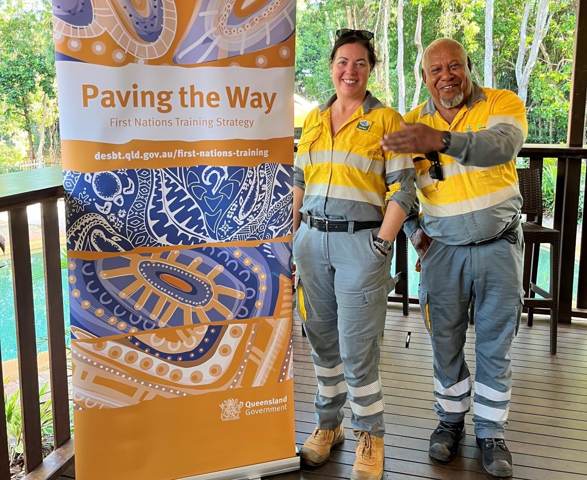 Tommy Sebasio is a First Nations man in a electricians uniform. He is standing with a woman in the same yellow and grey uniform next to a pull up banner that reads "paving the way: First Nations training strategy".