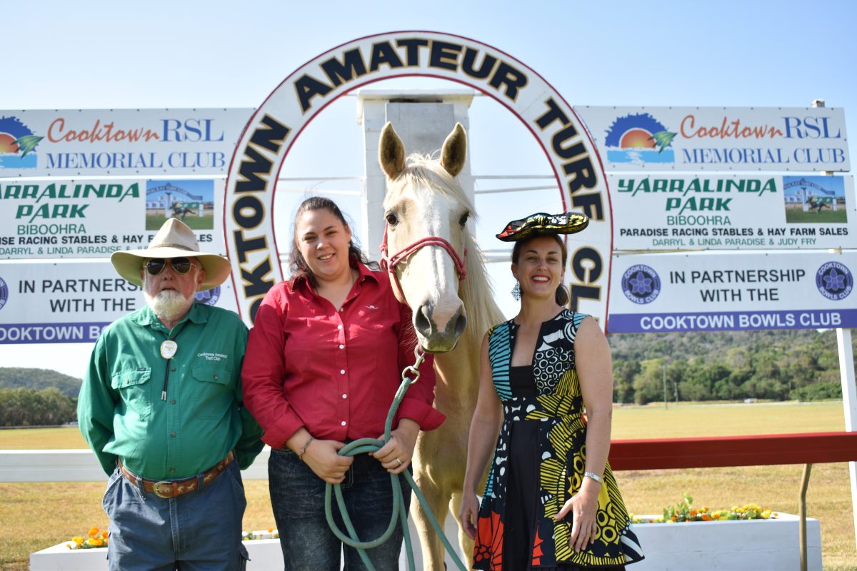 Three people and a horse stand in front of a round sign that reads "Cooktown Amateur Turf Club"
